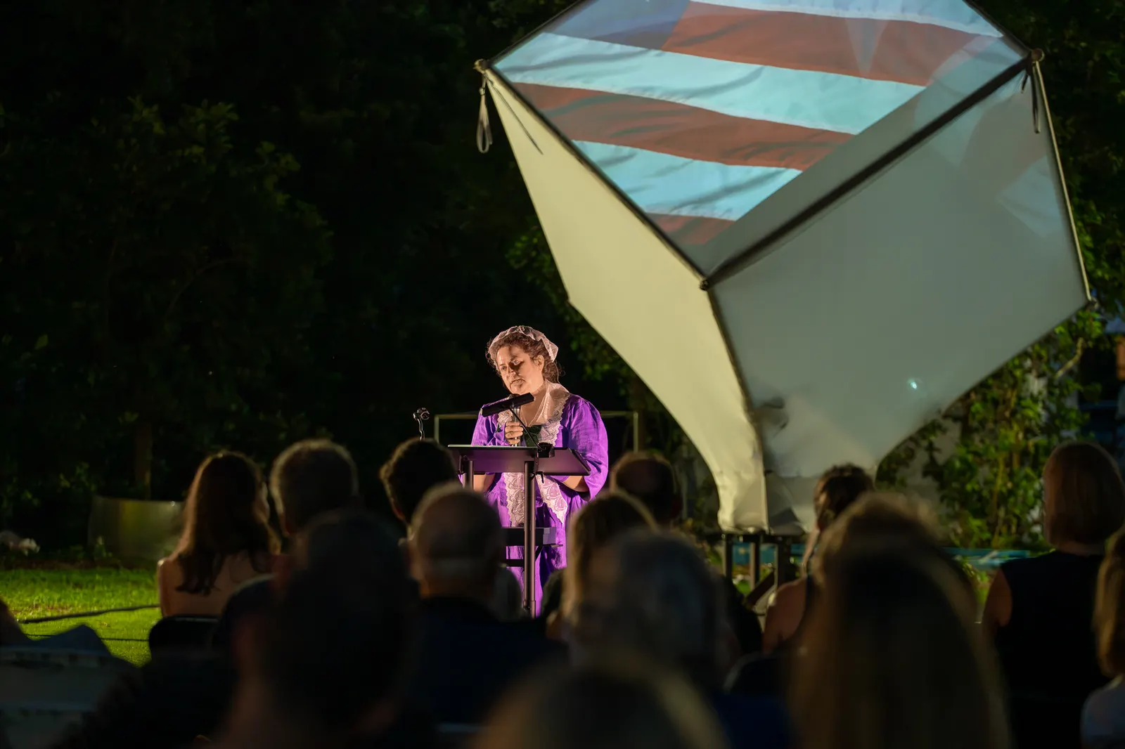 A woman in purple stands alone at the lectern before the striped cube.
