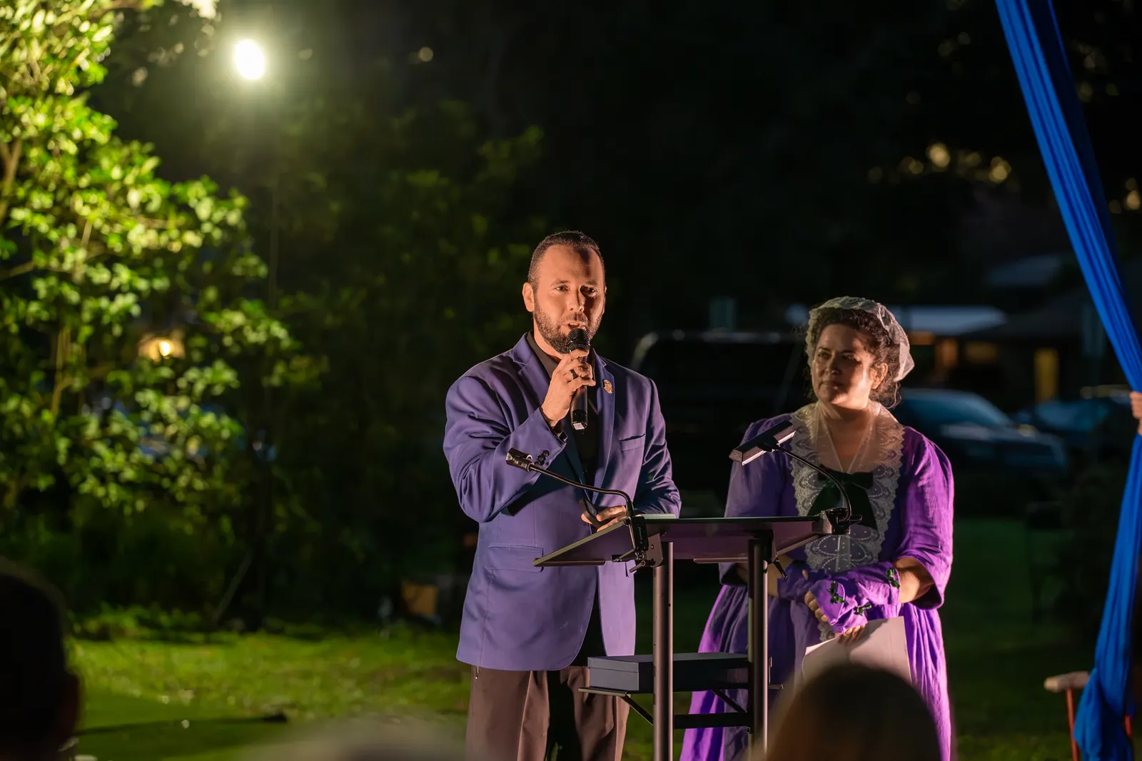 The man holds the microphone toward the older woman at the lectern.