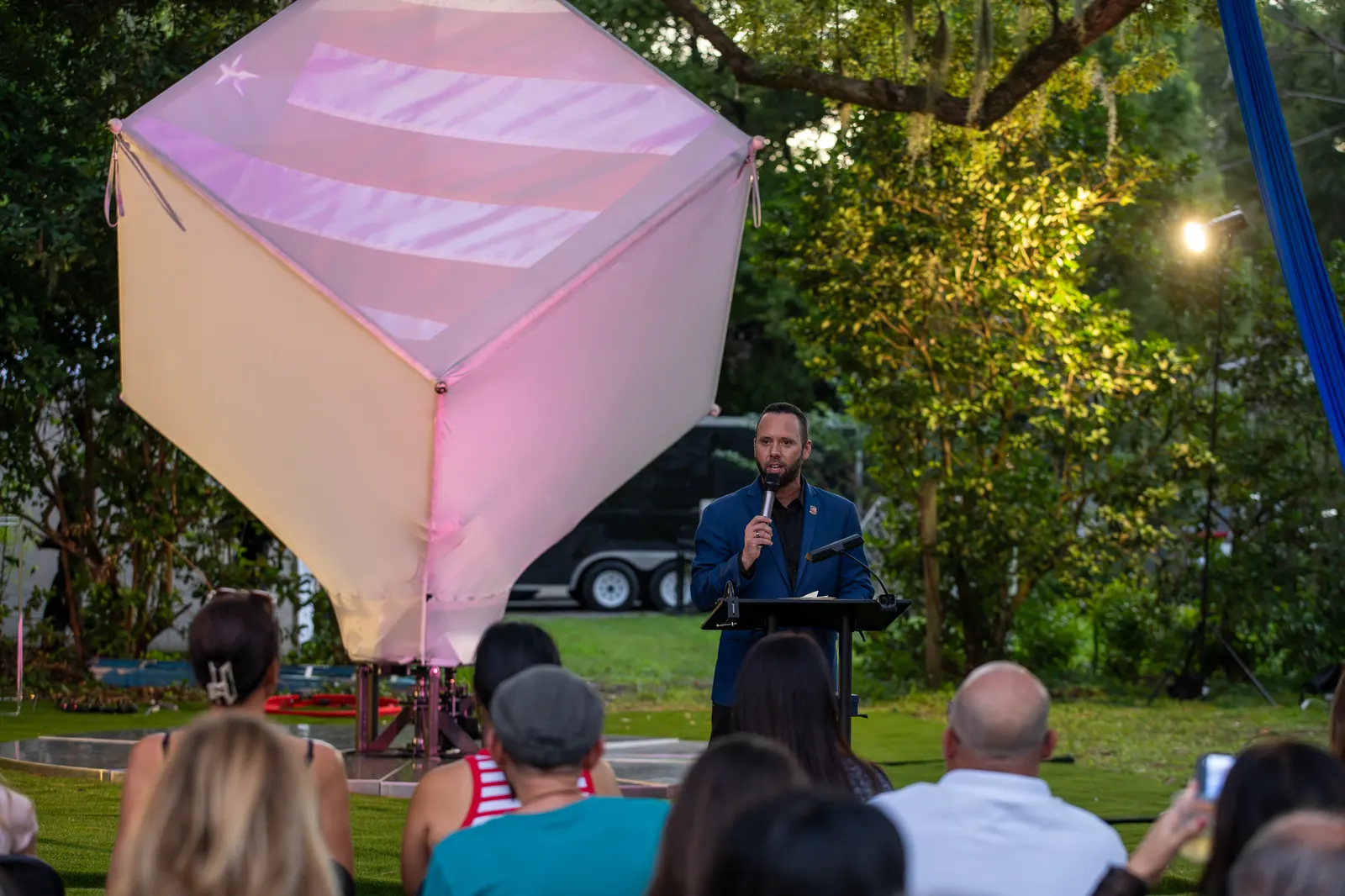 A speaker addresses the seated audience beside the glowing aerial cube.