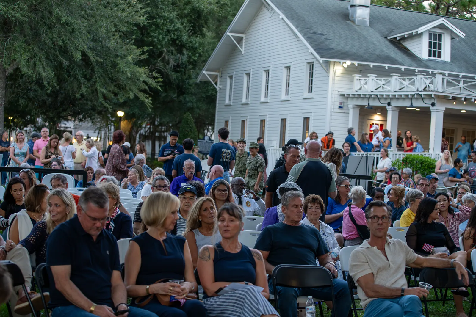 A seated audience fills the lawn in front of a white historic building.