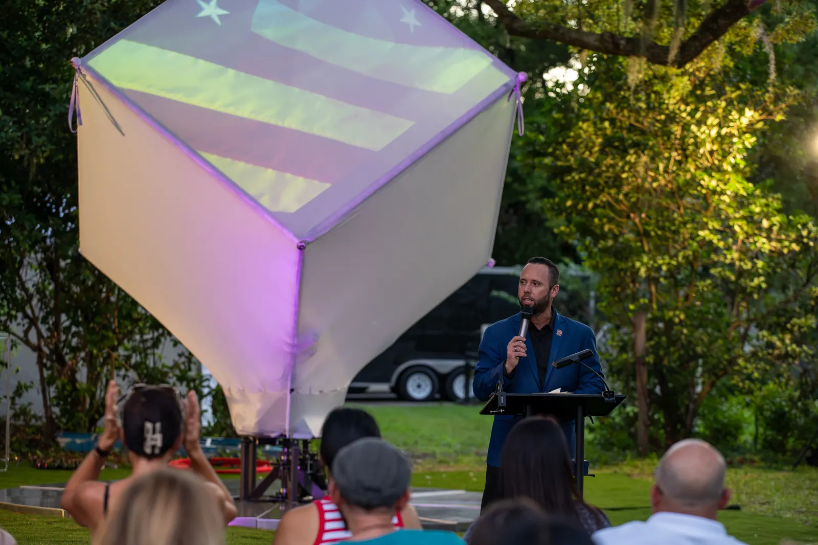 A man speaks from a lectern as colored light washes over the performance cube.