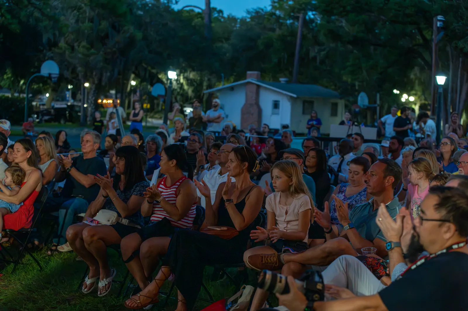 A large seated audience watches attentively in the blue evening light.