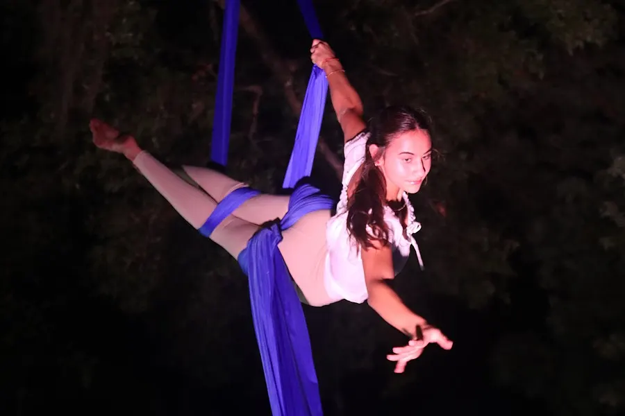 An aerial performer extends horizontally from blue silks under magenta light.