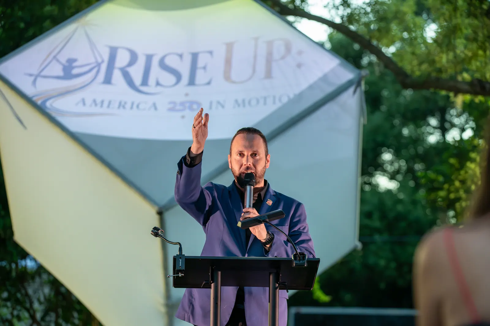 A man raises one hand while speaking before the Rise Up logo.