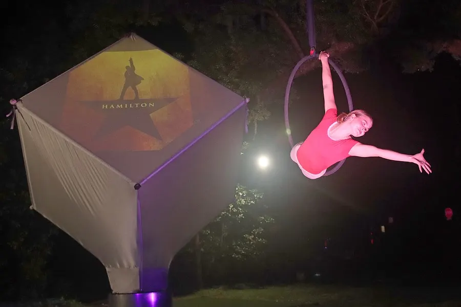An aerialist forms a compact inverted pose beside the glowing cube.