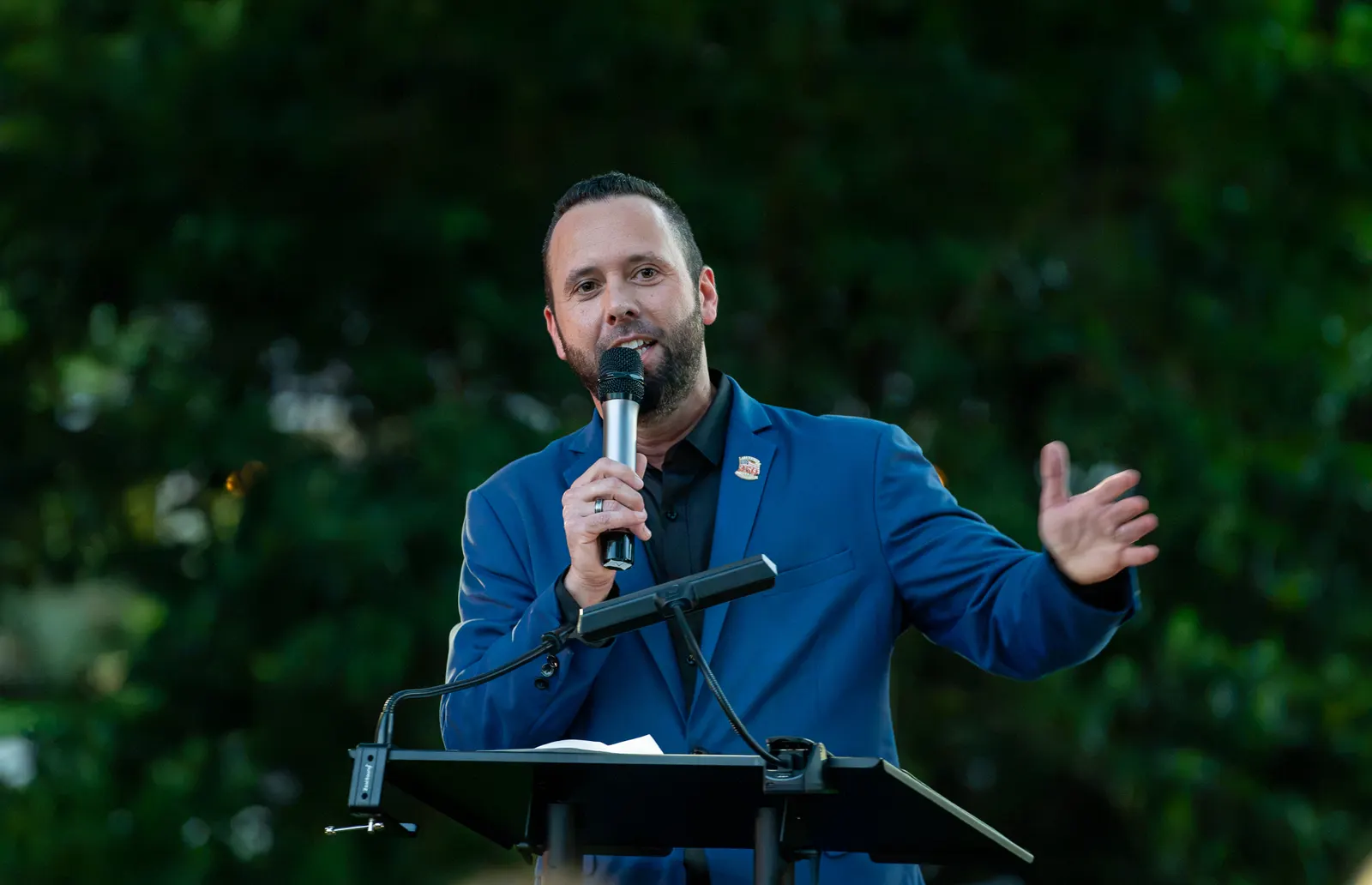 A bearded man speaks into a microphone at the outdoor lectern.