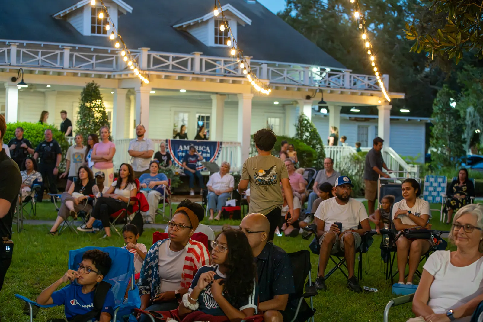 Families sit and stand near the white building as dusk deepens.