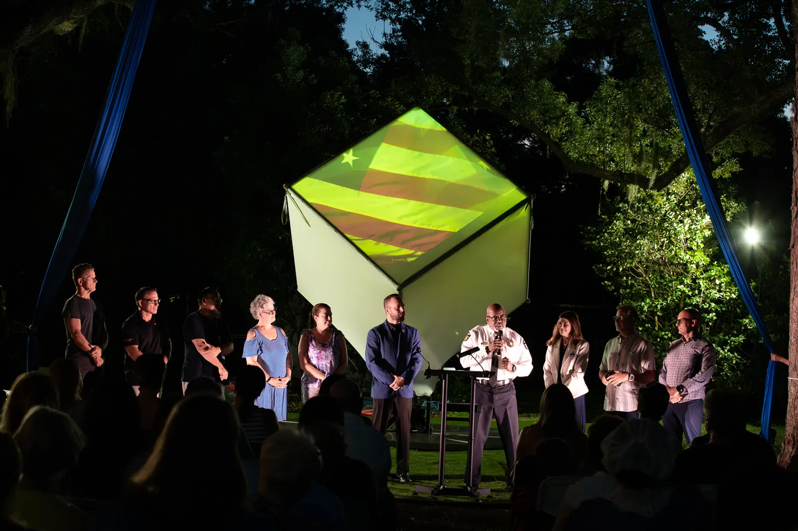 The uniformed speaker gestures as the group stands beneath the glowing cube.