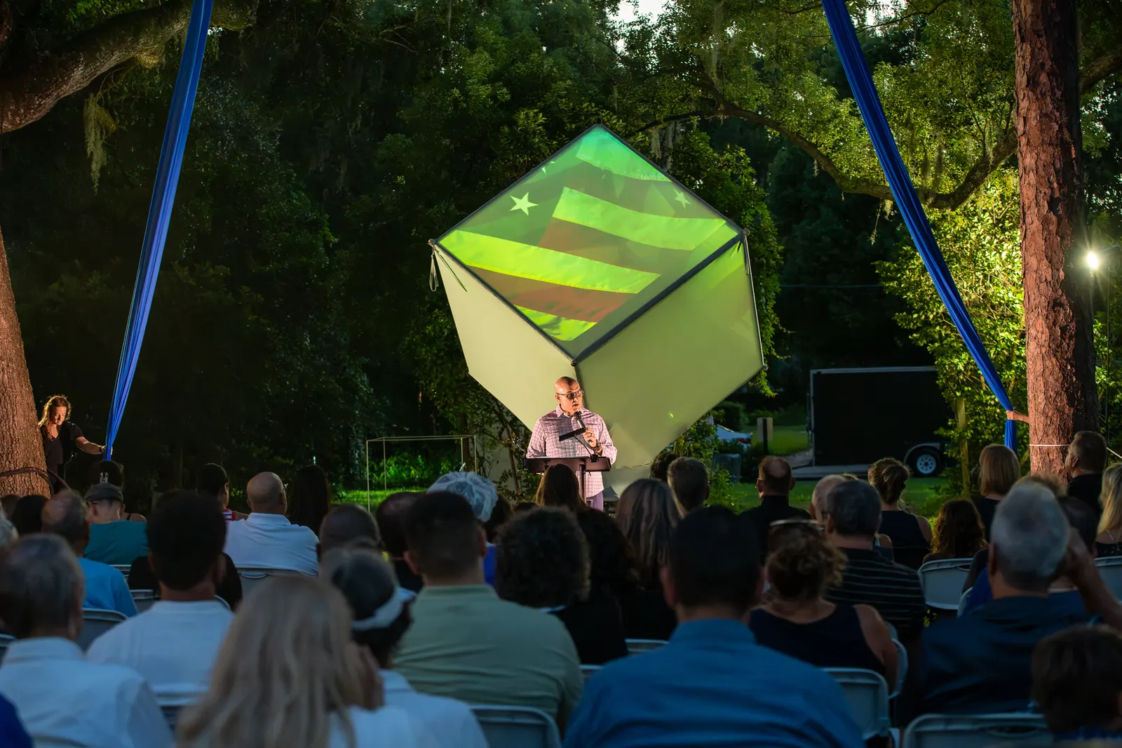 A speaker stands before rows of guests beneath the blue aerial rigging.