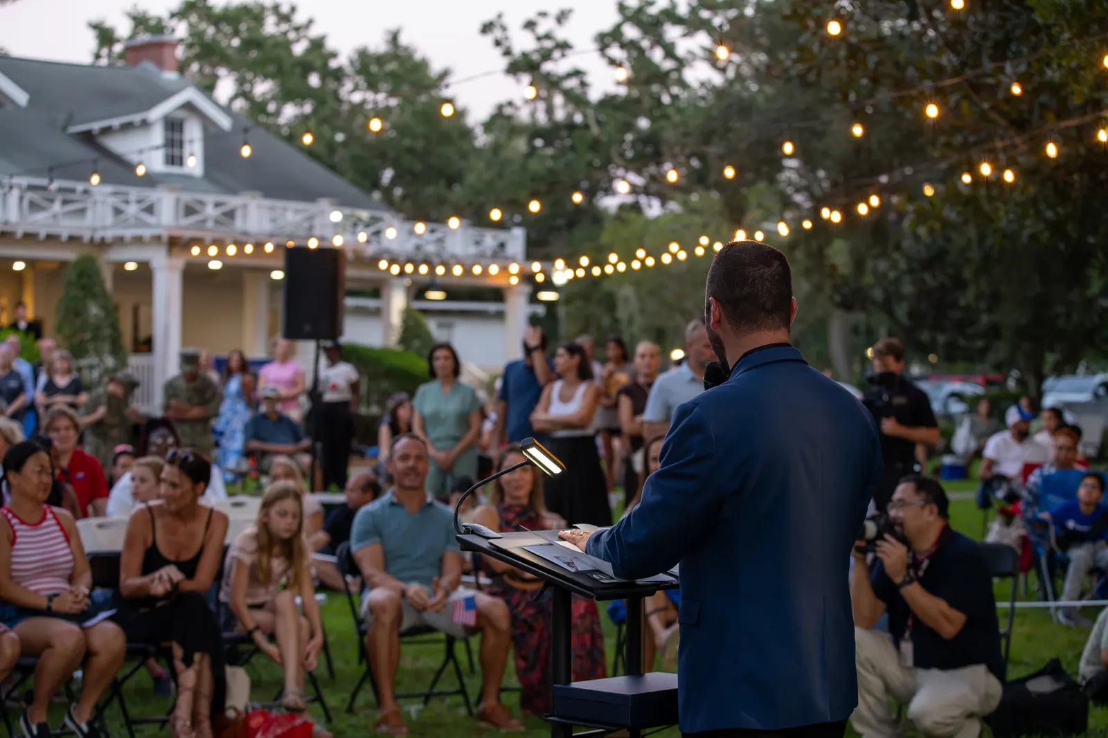 A speaker faces a large audience beneath strands of lights.