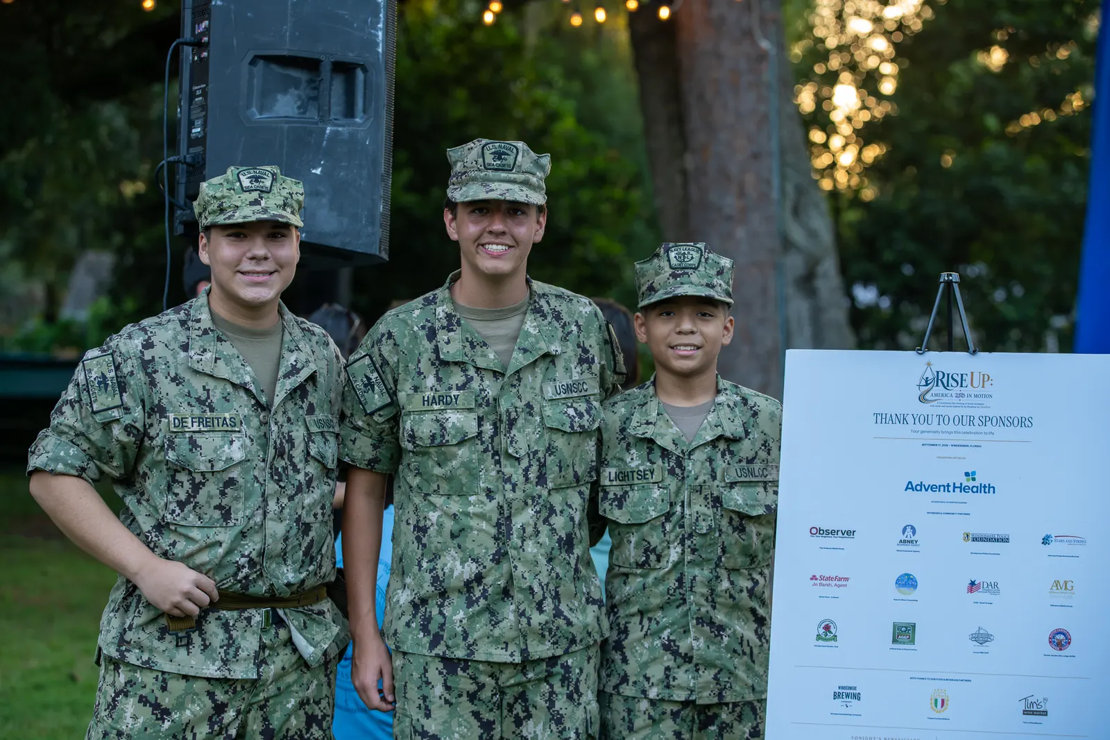 Two smiling Sea Cadets stand beside the event sponsor board.