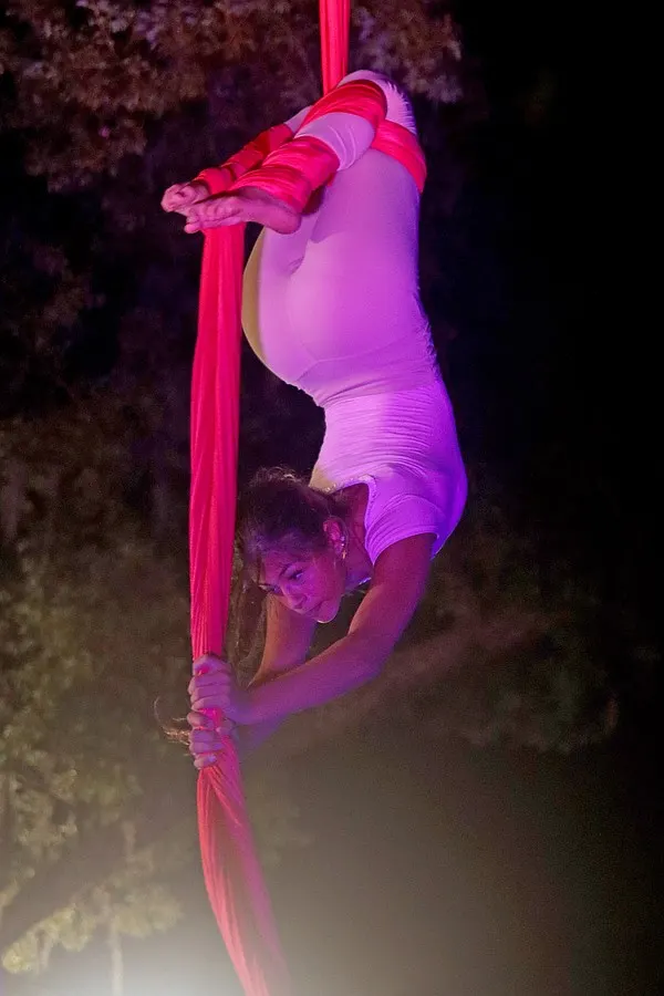 An aerialist climbs a red silk in a vertical nighttime portrait.