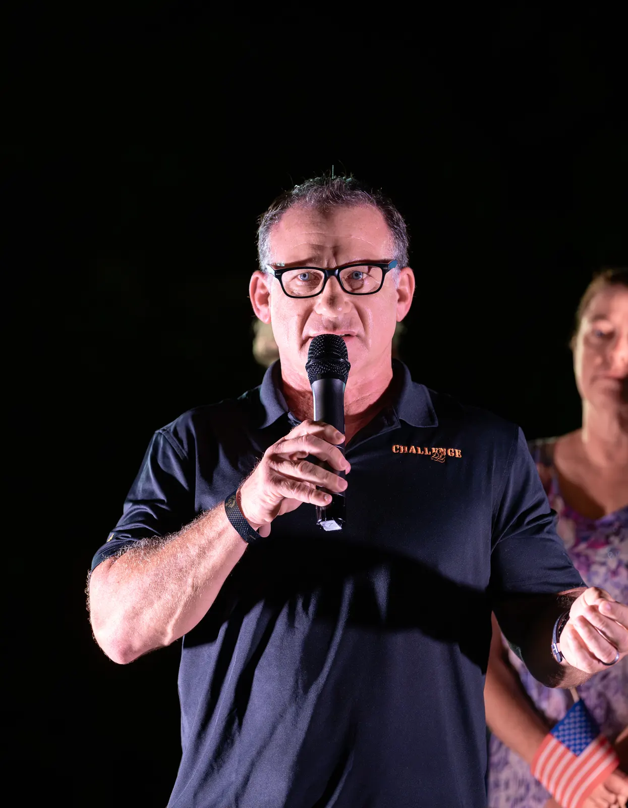 A man in a dark polo addresses the audience with a handheld microphone.