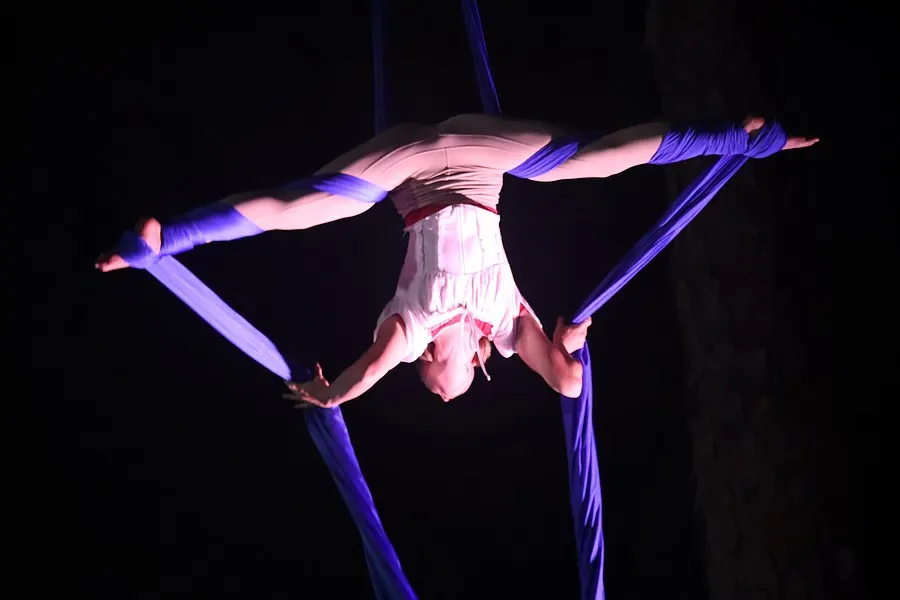 An aerialist holds a wide split high above the stage on blue silks.