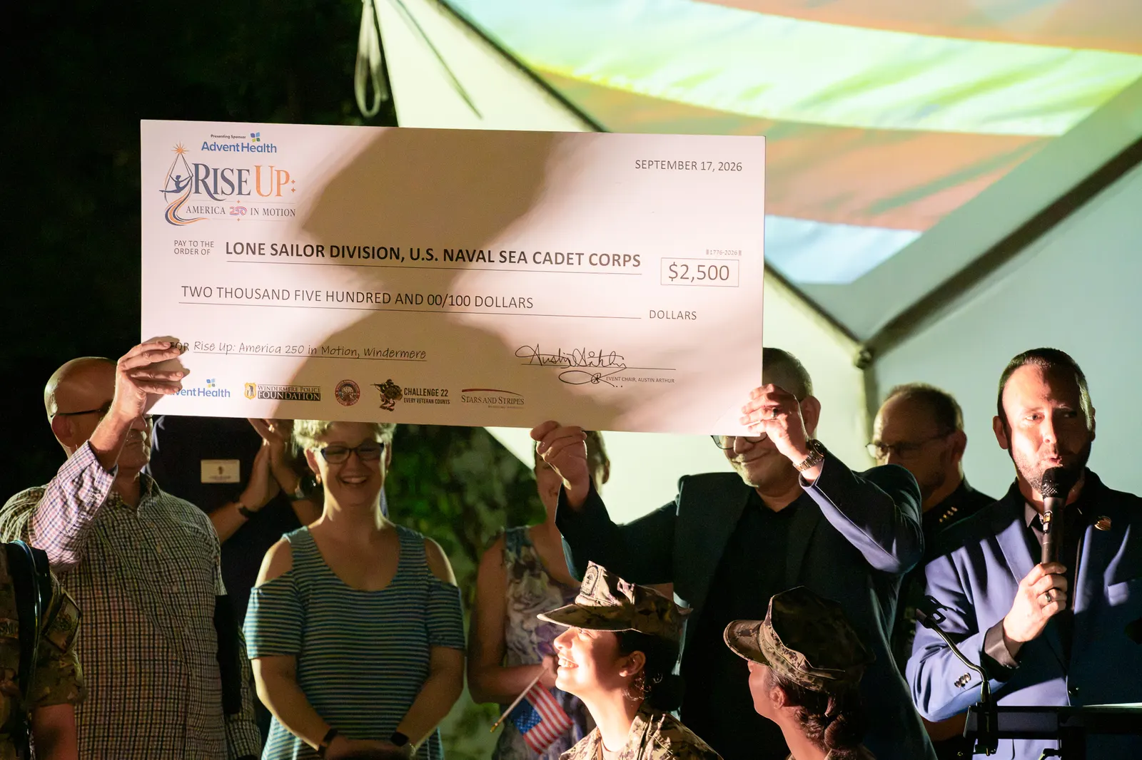 The large donation check is raised in front of the event logo.