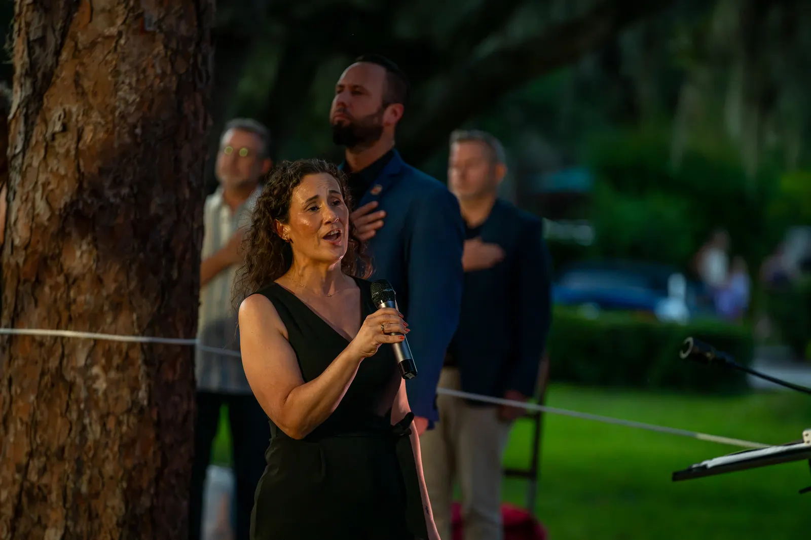 A woman sings beside a large tree as guests watch.