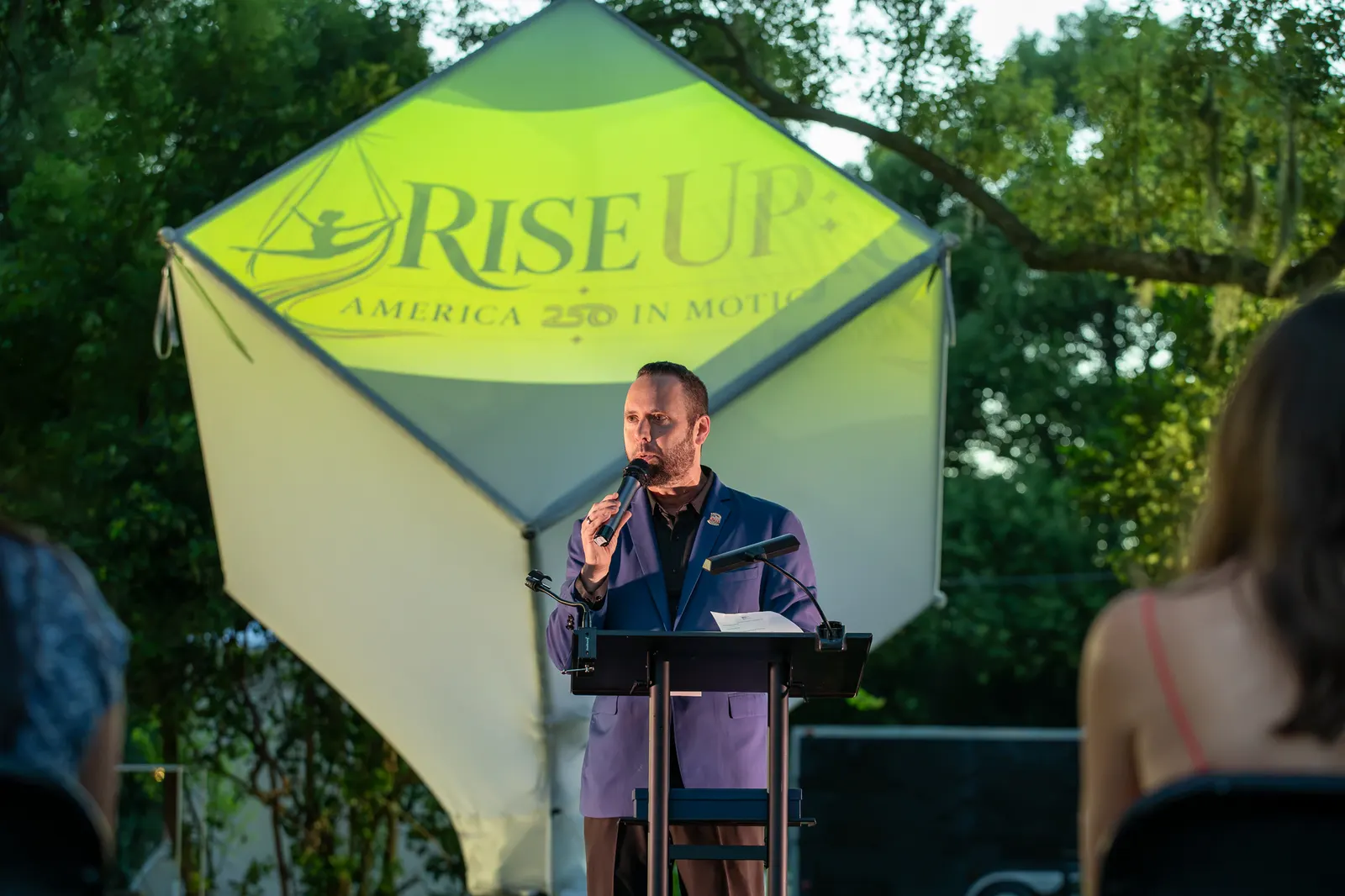 The speaker smiles at the microphone in front of the illuminated event logo.