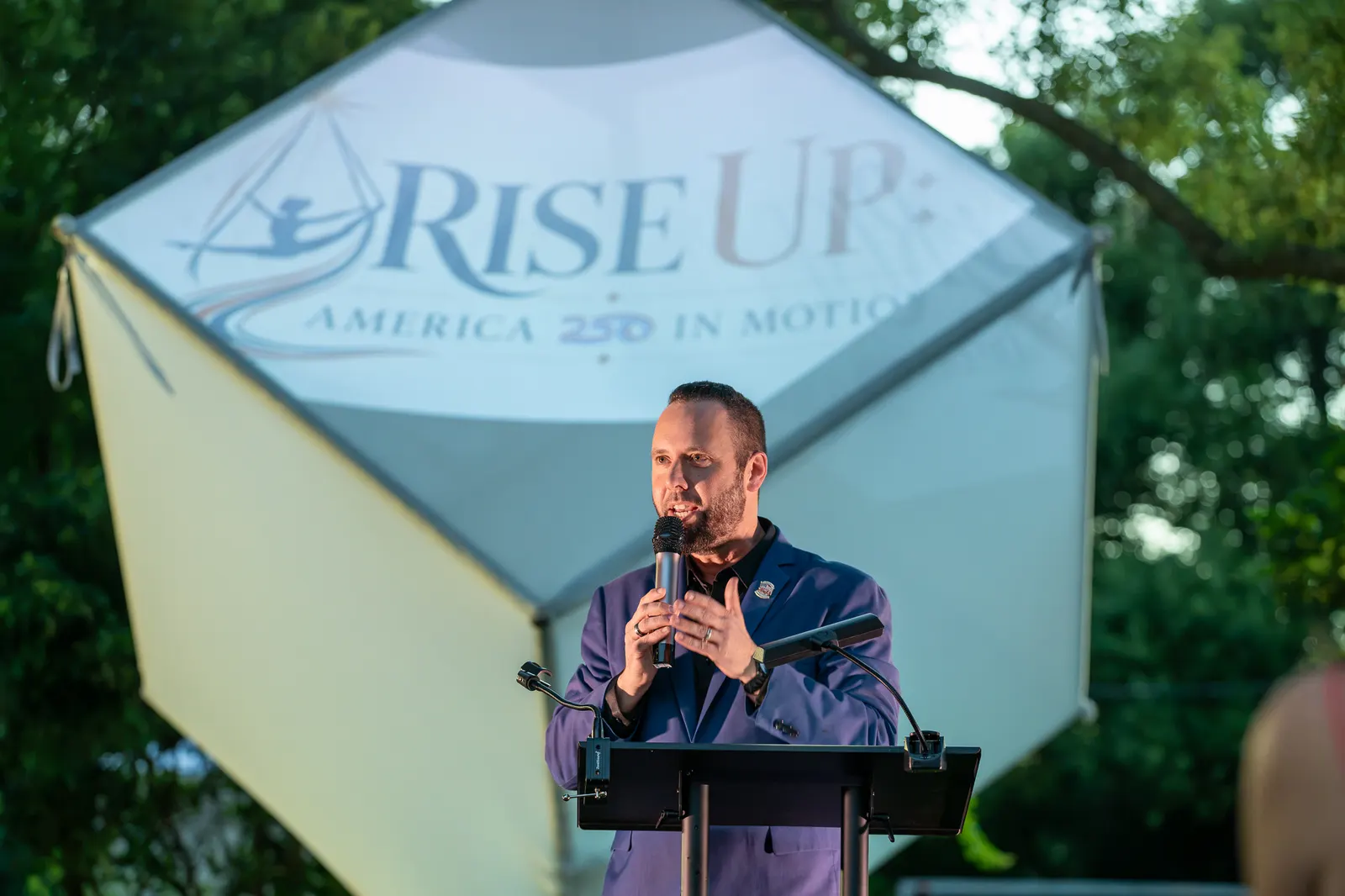 A man delivers remarks at the lectern with the Rise Up cube behind him.