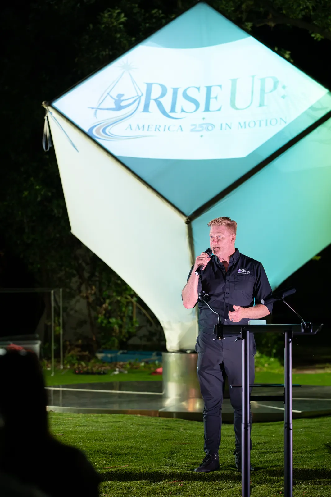 A vertical view frames the polo-shirted speaker before the Rise Up logo.